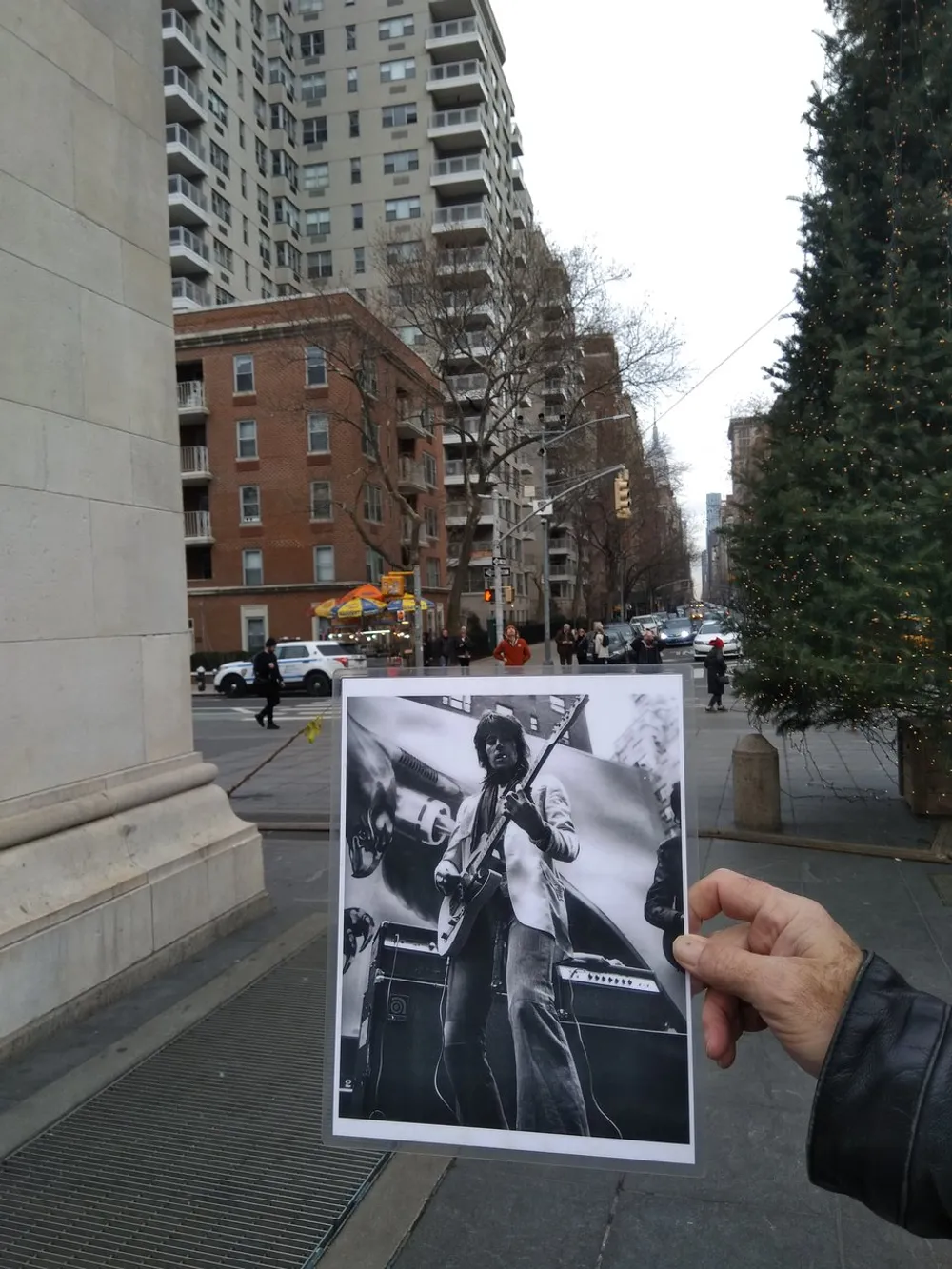 A person is holding up a black and white photograph of a guitarist in an urban setting aligning it creatively with the background street scene