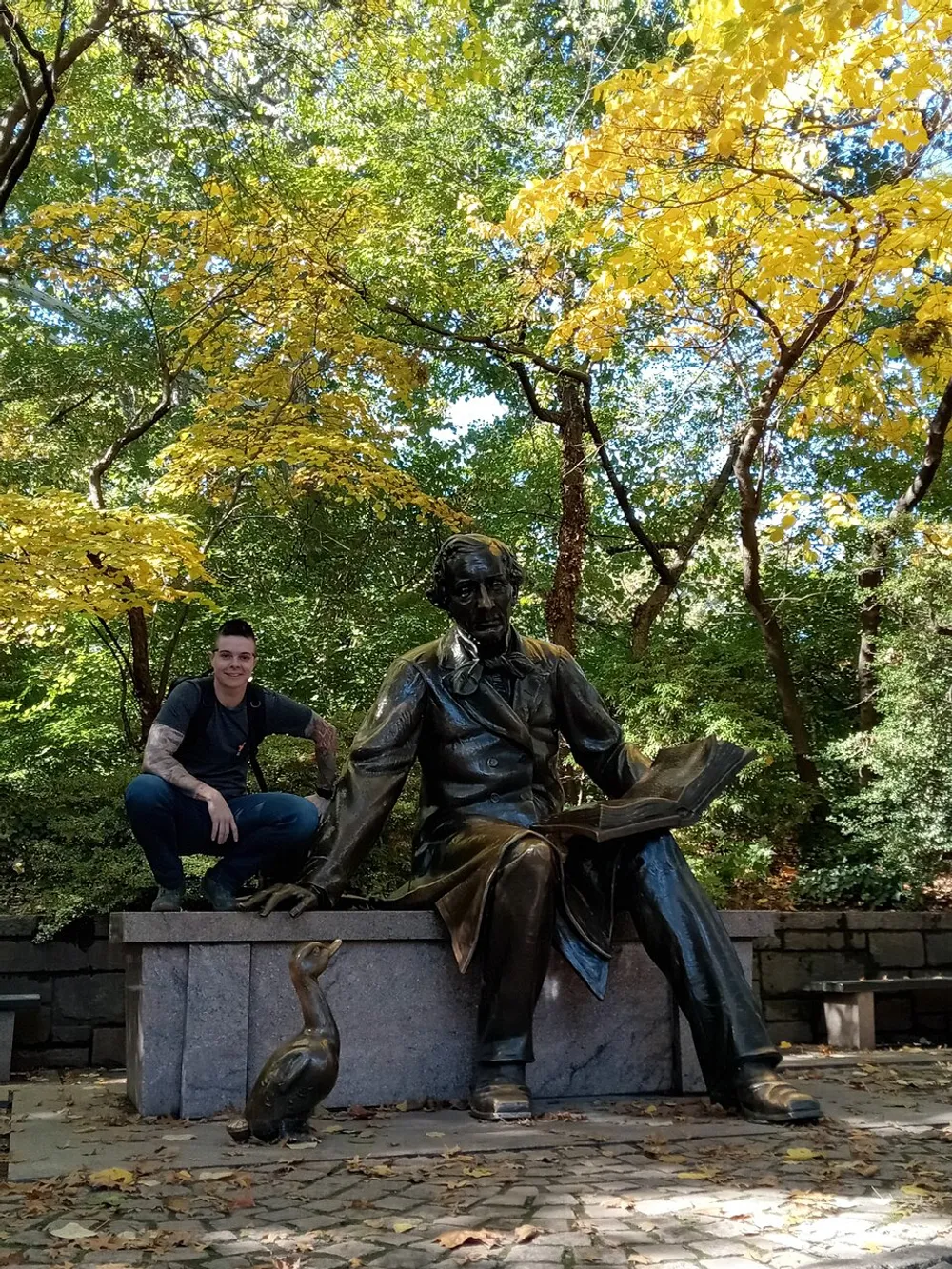 A person is posing next to a bronze statue of a seated figure holding a book with a duck at its base set against a backdrop of trees with autumn leaves