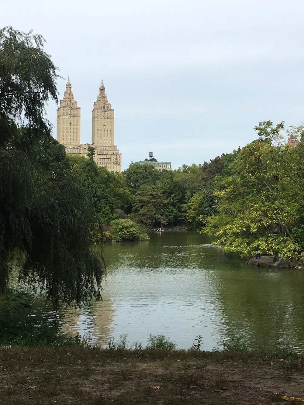The image shows a calm pond surrounded by greenery with the iconic twin-towered facade of the San Remo apartments rising in the background likely taken from within Central Park New York City