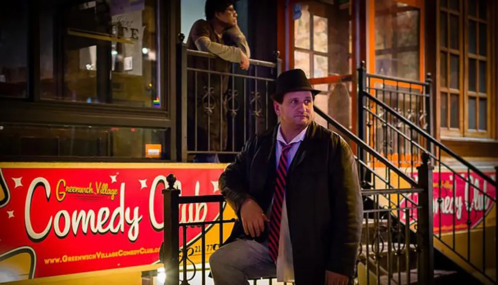 A man in a jacket and hat is seated on a metal staircase in front of the Greenwich Village Comedy Club at night