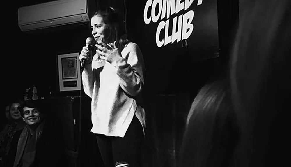A comedian performs on stage at a comedy club as the audience in the foreground looks on enjoying the entertainment