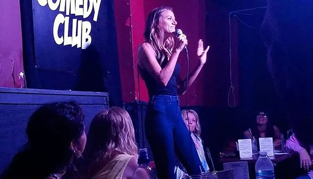A woman is performing stand-up comedy at a comedy club with an audience seated in the foreground