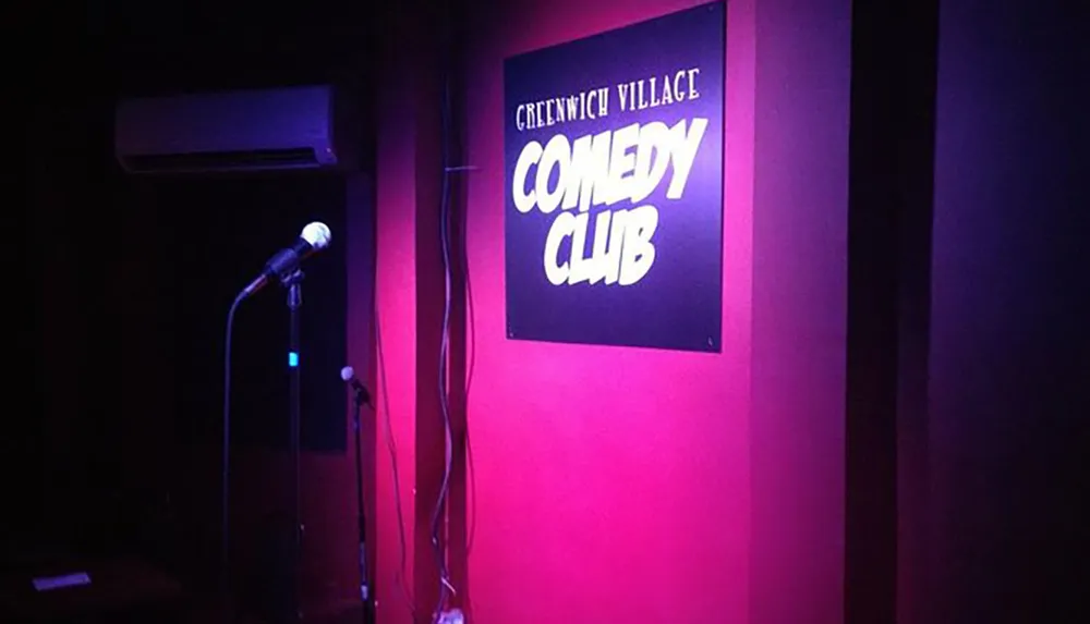 An empty stage with a microphone awaits a performer at the Greenwich Village Comedy Club highlighted by a purple hue and a solitary spotlight