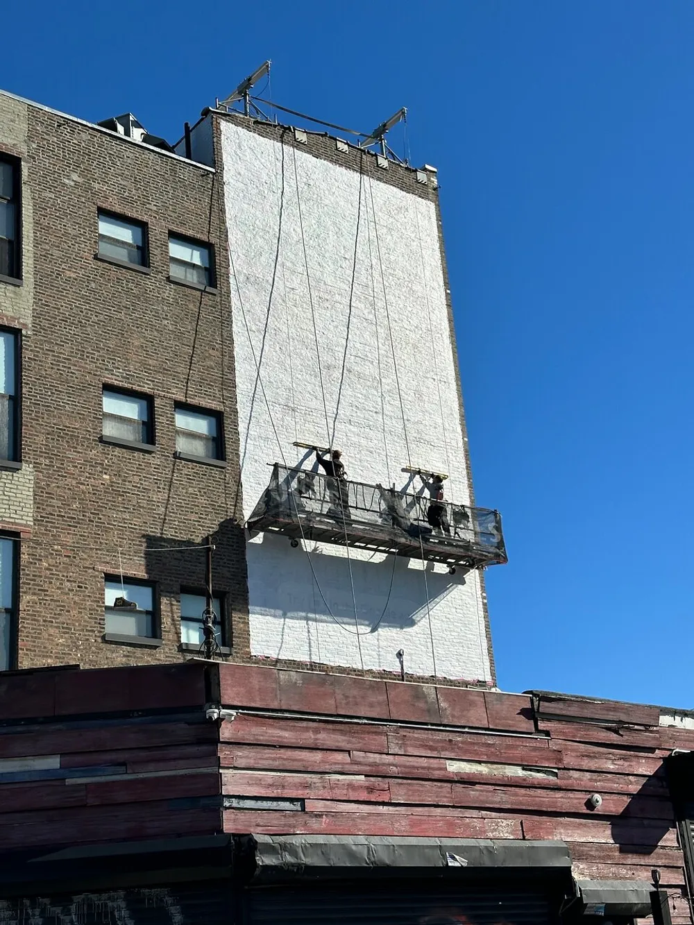 A suspended platform with workers is hanging on the side of a brick building possibly for maintenance or construction purposes against a blue sky background