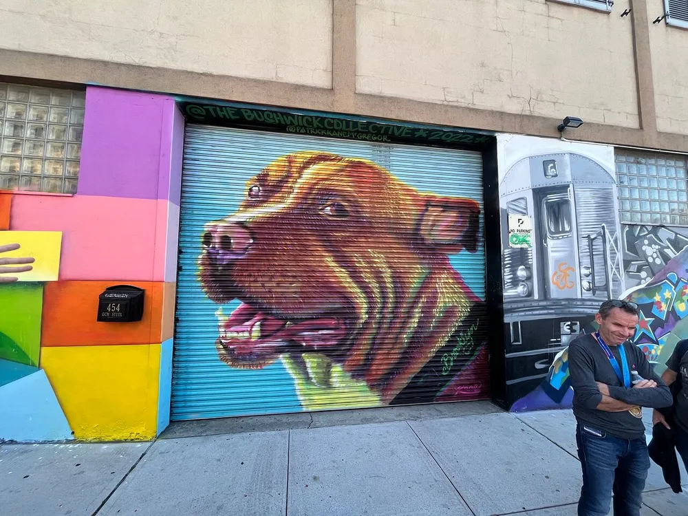 A colorful mural on a buildings roller shutter doors featuring a large dogs face to the left and a stylized subway train to the right with a smiling man standing in front
