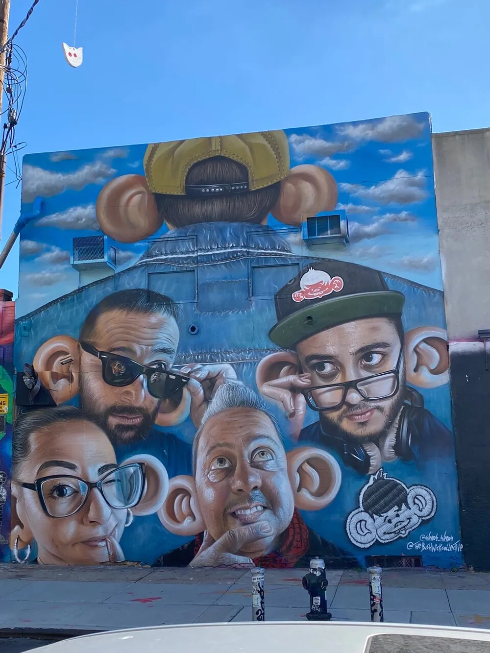 This image features a vibrant mural of four human faces with exaggerated ears and playful expressions along with a character wearing a cap viewed from the back set against a blue sky with clouds