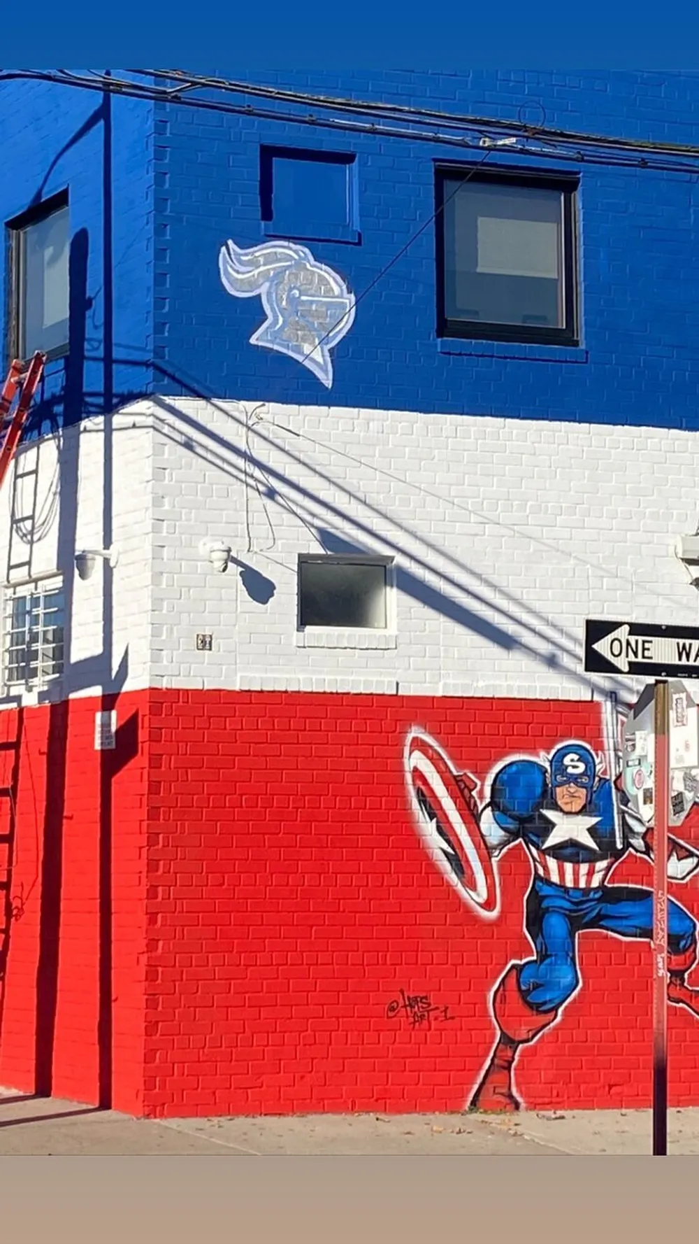 The image shows a vibrant street mural of a superhero character resembling Captain America on a red and blue painted building with a one-way street sign visible in the corner