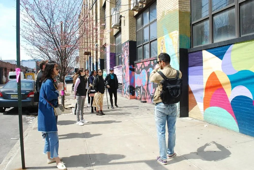 A group of people is attentively listening to a person facing them possibly a guide on a sunny day in front of a building with colorful street murals
