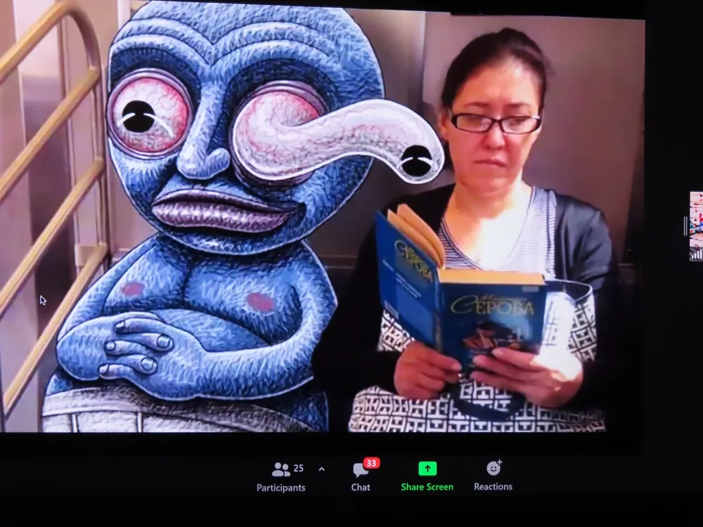 A screen during a video conference call displays a drawing of an alien character alongside a participant who is reading a book