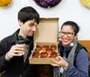 A man and a woman are smiling at the camera with the woman holding up a box of glazed pastries as the man holds a coffee cup both appearing cheerful and possibly celebrating a happy occasion or enjoying a casual treat
