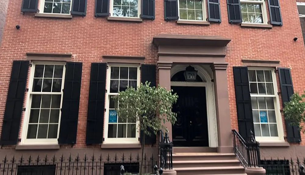 The image shows a traditional red-brick townhouse with black shutters a central black door and a small tree in front creating a classic urban residential scene