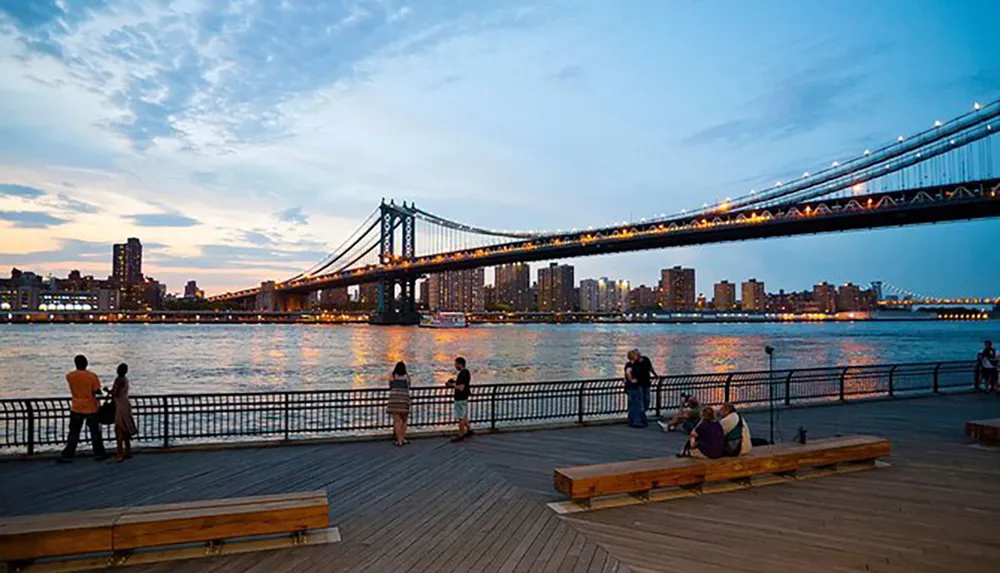 People enjoy the evening view of a lit-up suspension bridge from a waterfront boardwalk