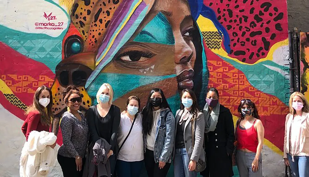A group of people wearing masks pose in front of a colorful mural featuring a large portrait of a womans face