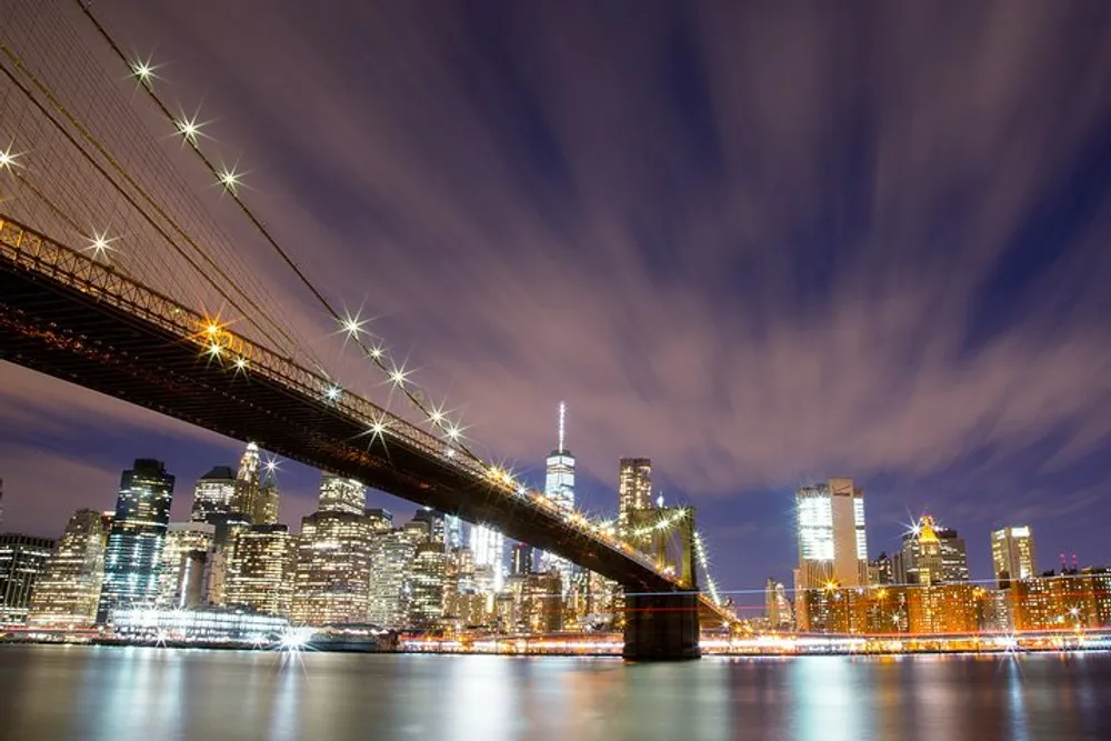 The image shows a long exposure night shot of the Brooklyn Bridge with radiant stars of light and streaky clouds above set against the backdrop of the illuminated New York City skyline