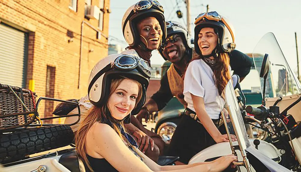 A group of four cheerful people wearing motorcycle helmets is posing with a vintage-style sidecar motorcycle on a sunny street