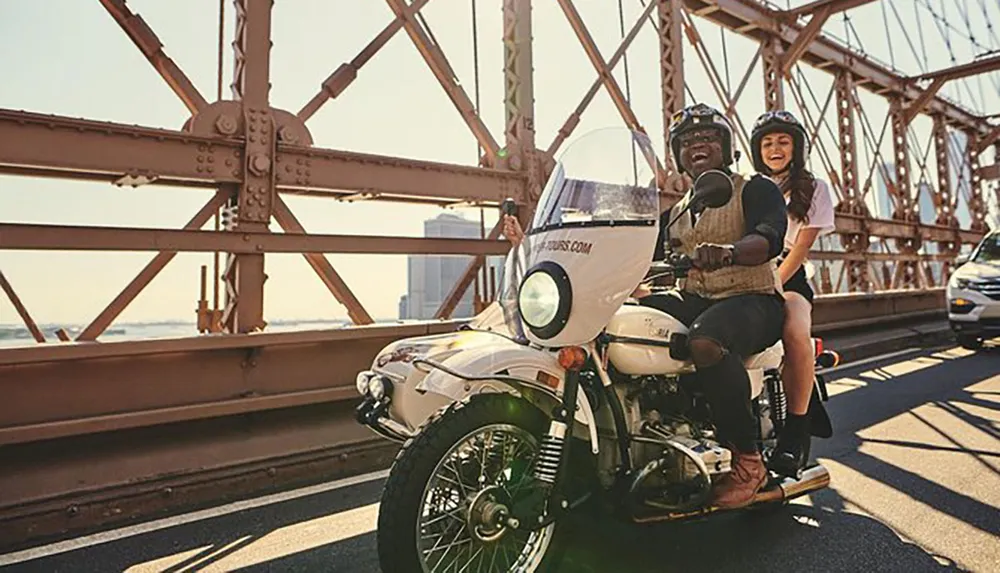 Two people are riding a motorcycle with a sidecar smiling and enjoying their time together on a sunny day crossing a bridge with steel structures visible around them