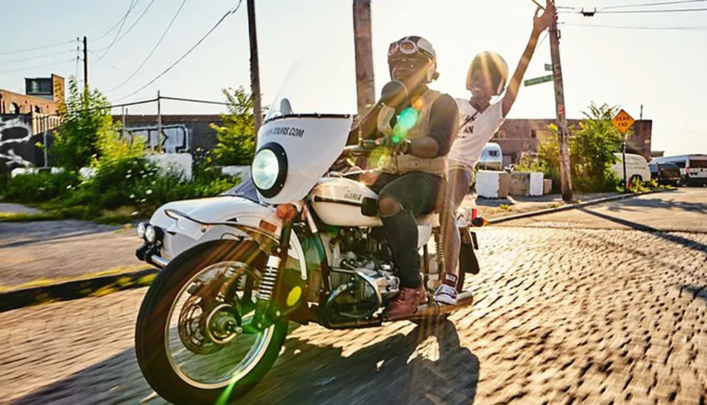 Two individuals are joyfully riding on a classic motorcycle with the passenger waving on a sunny day down an urban street lined with cobblestones and industrial buildings