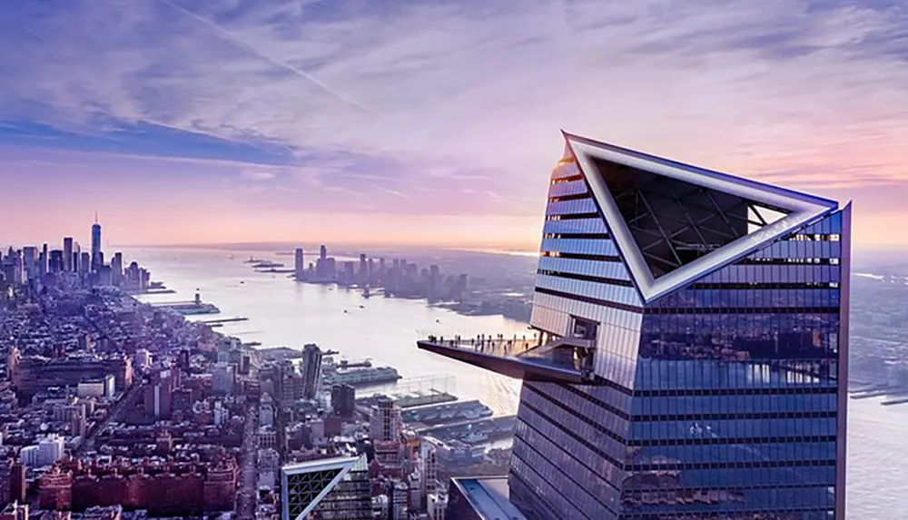The image shows a modern skyscraper with a distinctive angled top overlooking a panoramic view of a citys skyline during what appears to be dawn or dusk with a river and other tall buildings in the background
