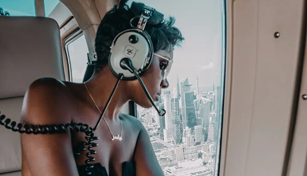 A person wearing headphones is looking contemplatively out of a helicopter window at a skyline of skyscrapers