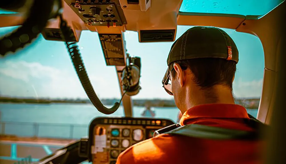 A person in a cap and sunglasses is piloting a seaplane with a view of the cockpit instruments and a water landscape outside