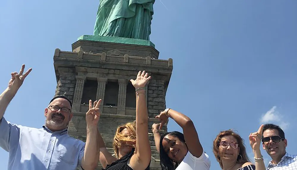 A group of people are posing joyfully in front of the Statue of Liberty with some of them raising their hands in a peace sign gesture