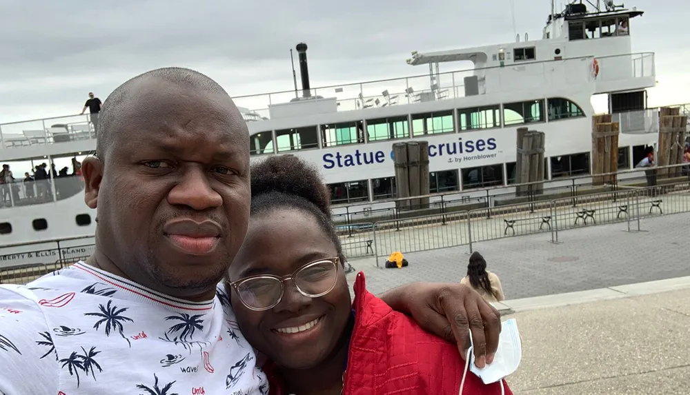 Two people are taking a smiling selfie with a Statue Cruises ferry in the background