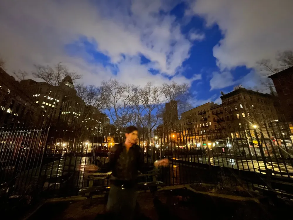 A blurred figure walks through a park at dusk with city lights and buildings in the background under a dramatically clouded evening sky