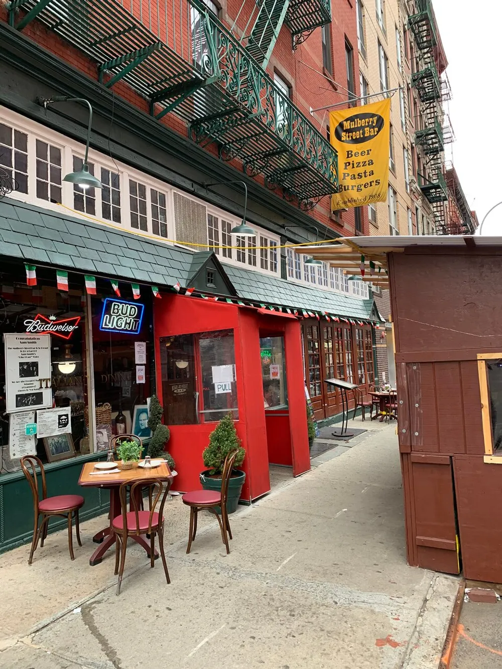 The image shows a quaint street view with the Mulberry Street Bar featuring outdoor tables and classic Italian restaurant signage nestled among traditional-style buildings with iron fire escapes