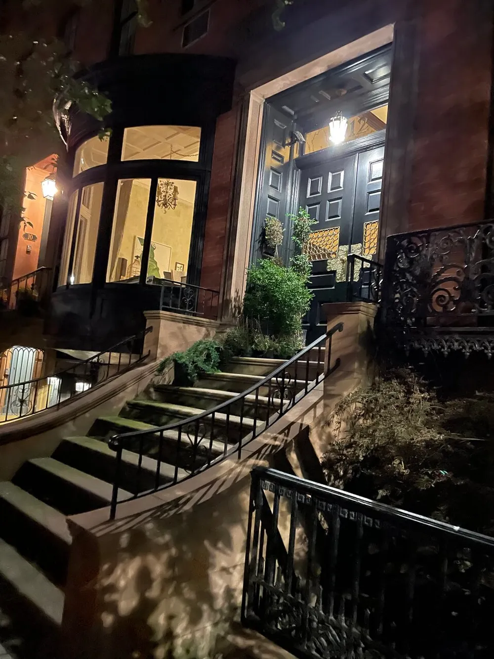 The image shows a well-lit entrance to a traditional brownstone at nighttime with steps leading up to a dark wooden door framed by decorative elements and surrounded by potted plants