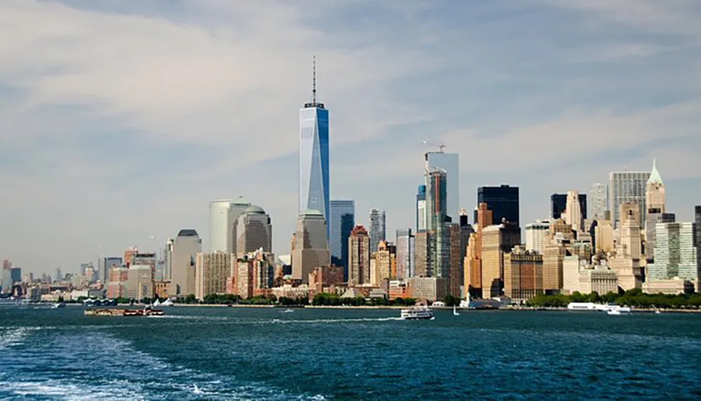 The image shows a daytime view of the Manhattan skyline with the One World Trade Center standing prominently against a clear sky as seen from across the water with boats on the surface