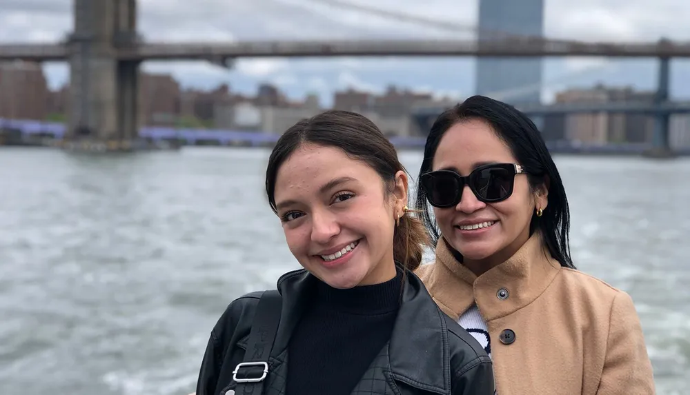 Two smiling individuals pose for a photo with a prominent bridge in the background likely taken on a boat or waterfront