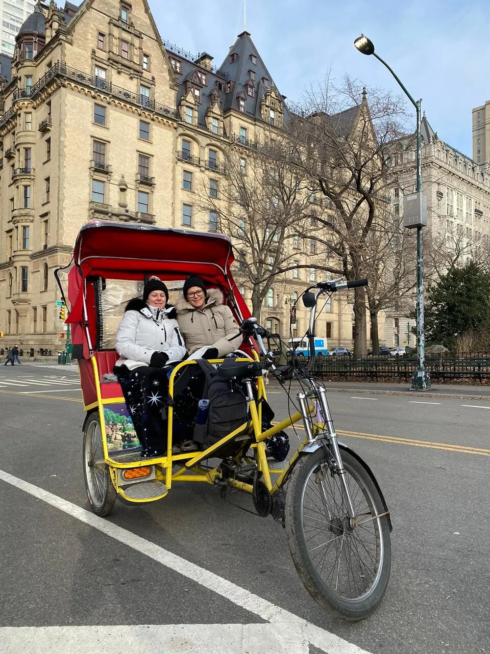 Two individuals are smiling for a photo while seated in a red and yellow pedicab in front of a historic building with leafless trees and a streetlamp in the background