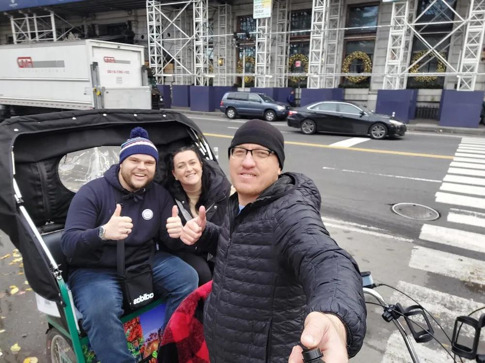 Three people are smiling and giving thumbs-up from a pedicab on a city street with one of them taking a selfie