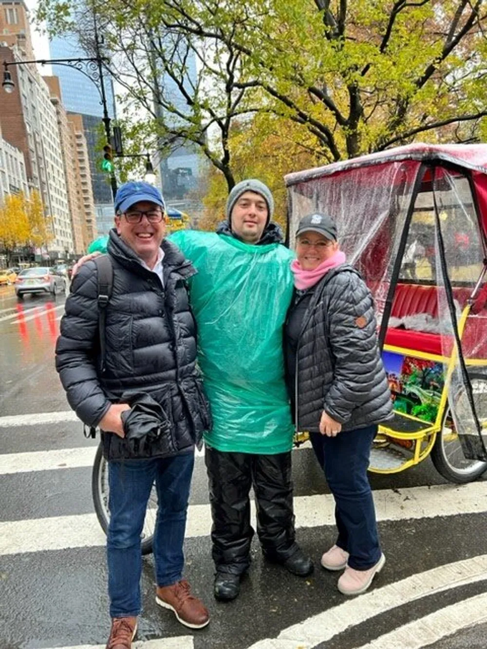 Three people are smiling for the camera on a city street with a rickshaw in the background and the person in the middle is wearing a green rain poncho