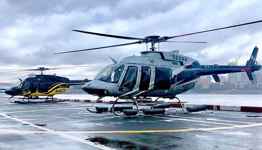 A pair of helicopters is parked on a helipad with one featuring a yellow and black livery and the other in a darker color scheme against an overcast sky with a city skyline and bridge faintly visible in the background