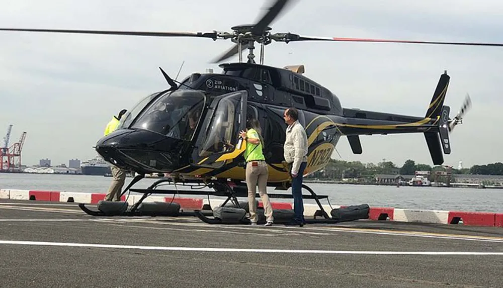 A person is exiting a helicopter on a helipad near a body of water while another individual in a reflective vest assists