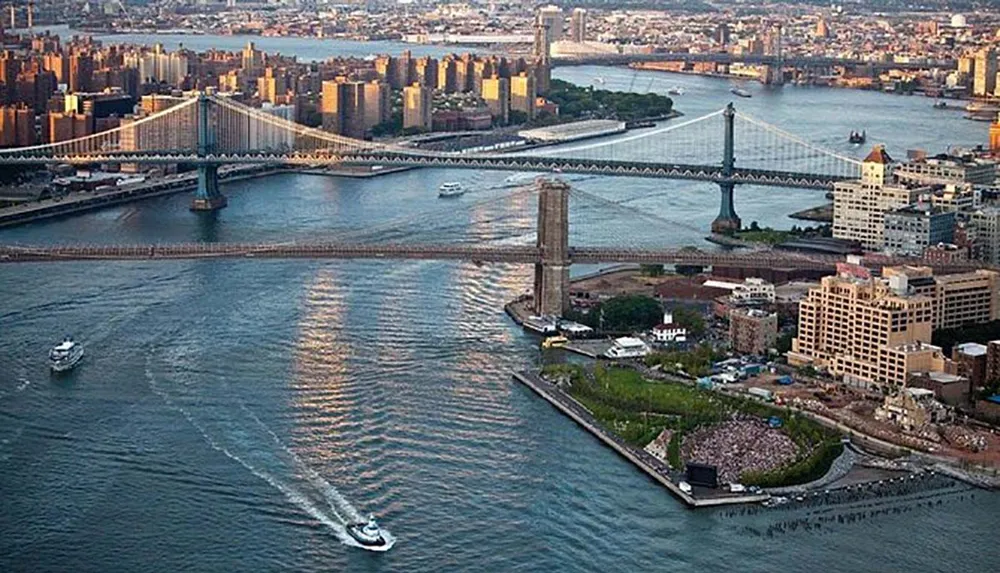 The image shows an aerial view of the East River with the Manhattan and Brooklyn Bridges connecting Manhattan to Brooklyn and a boat creating ripples on the water as the sun begins to set