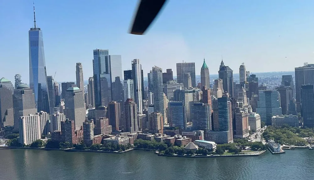 The image shows a sweeping aerial view of a densely packed urban skyline with skyscrapers likely captured from a helicopter featuring a prominent slender tall building which appears to be One World Trade Center in New York City