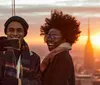 A smiling man and woman are taking a selfie with a smartphone against a backdrop of a city skyline at sunset