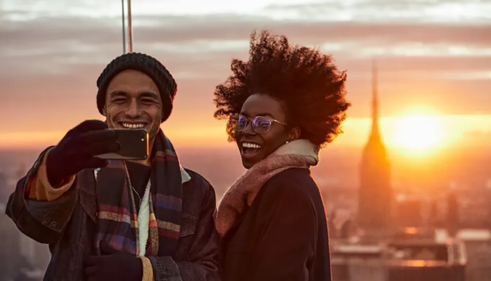 A smiling man and woman are taking a selfie with a smartphone against a backdrop of a city skyline at sunset