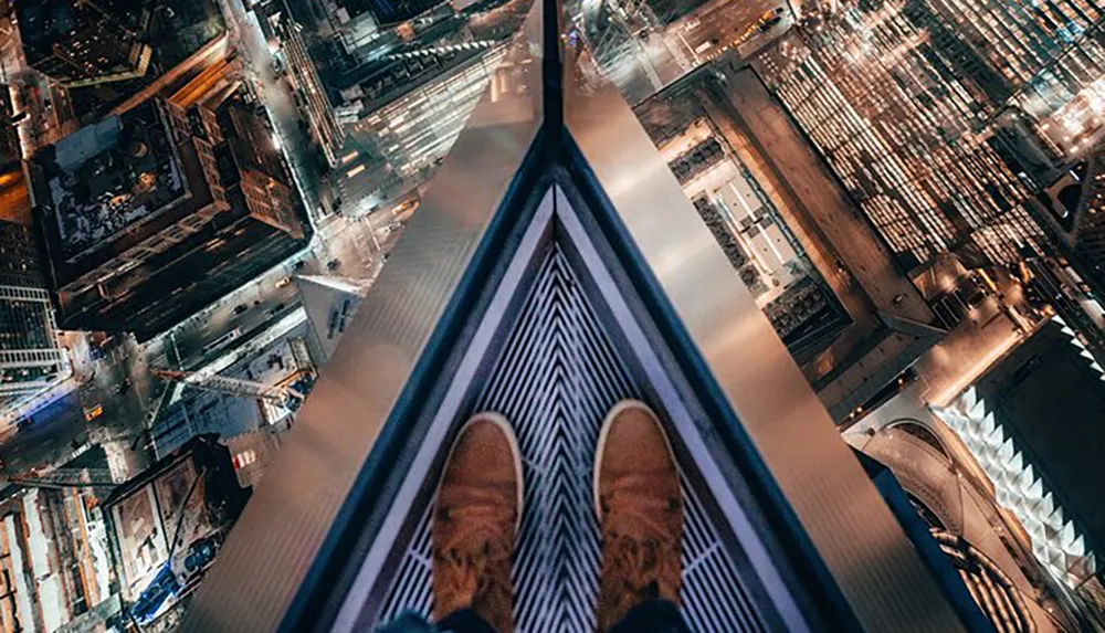 The image shows a persons feet standing at the edge of a high skyscraper providing a dizzying perspective of the illuminated city streets far below