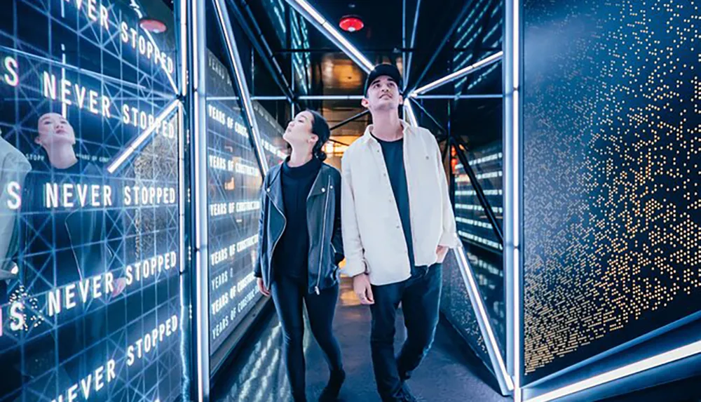A man and a woman are walking through a modern illuminated tunnel with dynamic light patterns and text displays looking up at the vibrant designs