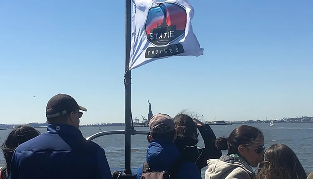Passengers on a boat are looking towards the Statue of Liberty with a flag reading STATE CRUISES fluttering in the foreground