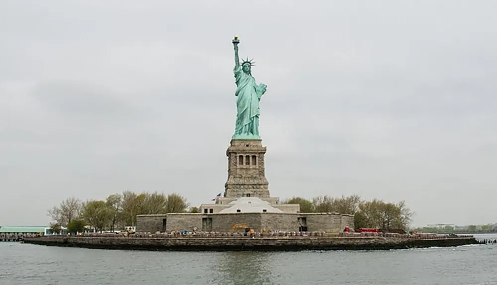 The image shows the Statue of Liberty on its pedestal situated on an island with overcast skies in the background