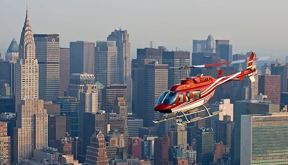 A red helicopter is flying over a dense urban skyline with skyscrapers including the iconic Chrysler Building