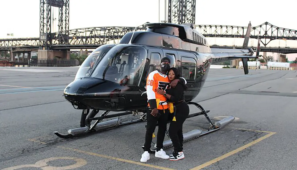 Two individuals pose for a photo in front of a black helicopter with a lift bridge in the background