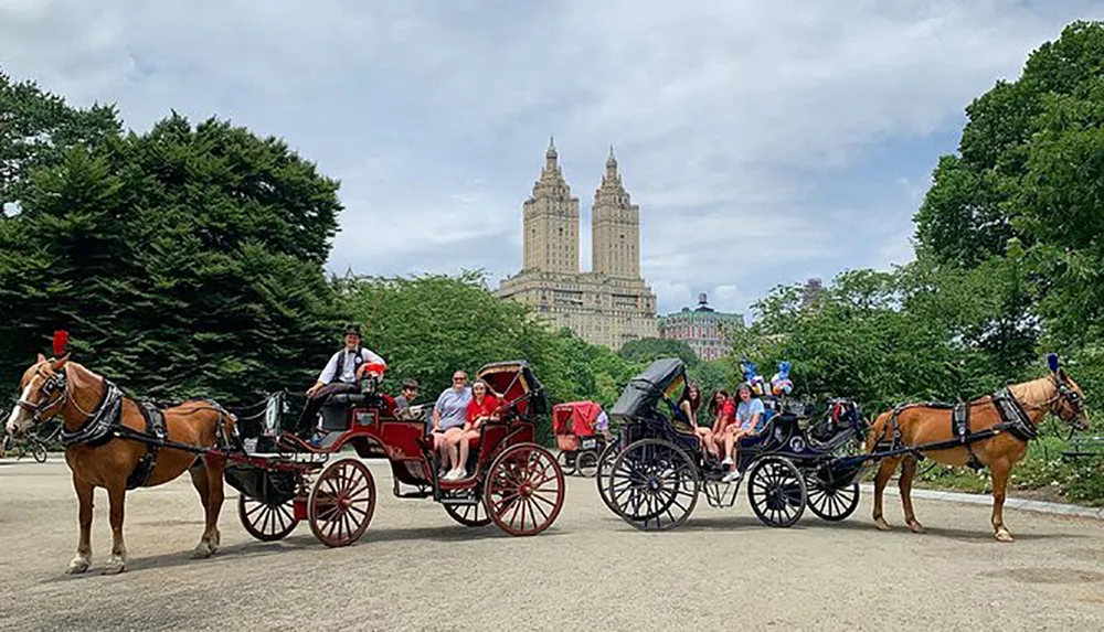 Two horse-drawn carriages carry passengers through a park with a backdrop of towering buildings