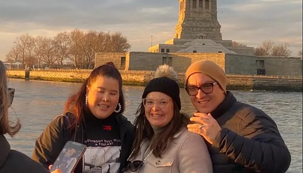 Three people are smiling for a photo with the Statue of Liberty in the background during what appears to be sunset