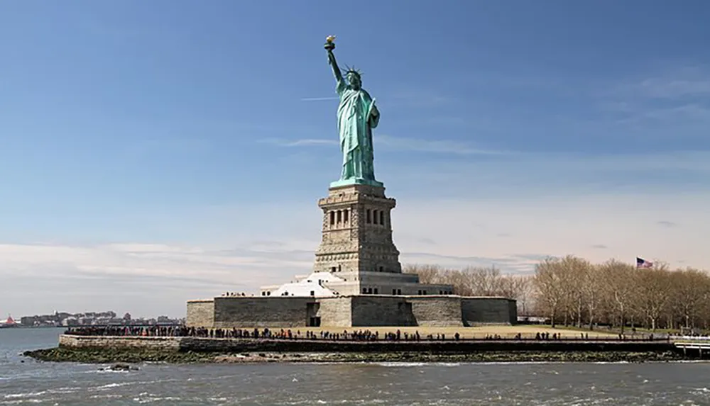 This image shows the Statue of Liberty against a blue sky with scattered clouds on its pedestal on Liberty Island surrounded by visitors