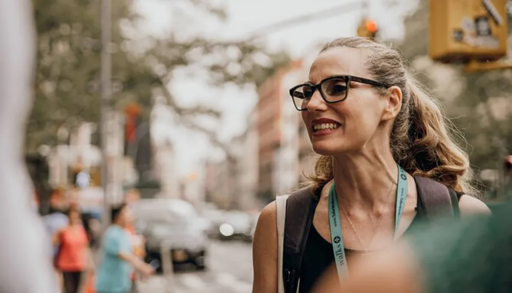 The image shows a smiling woman with glasses looking to her right on a busy city street with blurred pedestrians and traffic in the background